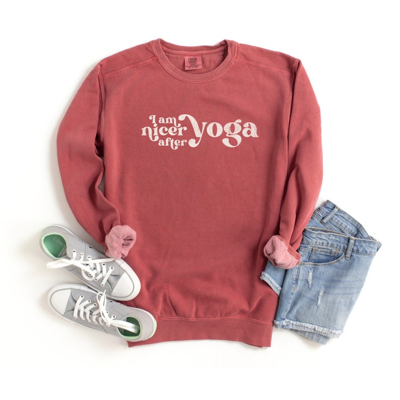 May include: A maroon sweatshirt with the text "I am nicer after yoga" in white. The sweatshirt is laid flat on a white surface with a pair of gray sneakers and a pair of blue denim shorts.