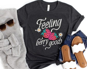 May include: Dark gray t-shirt with the words "Feeling berry good" in white script, featuring a strawberry graphic. The shirt is styled with sunglasses, a gray sweater, denim shorts, and white sandals.