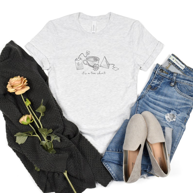 May include: A heather grey t-shirt with a graphic of tea bags, a teacup, and a tea pyramid. The text "it's a tea shirt" is printed below the graphic.