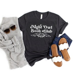 May include: A dark gray t-shirt with the text "Night Owl Book Club" in white with a vintage owl graphic. The shirt is laying on a white surface with a gray sweater, blue jeans, and white sandals.