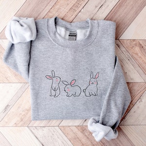May include: A grey sweatshirt featuring a graphic of three bunnies in a row. The bunnies are drawn in a simple, cartoon style with pink noses and ears.