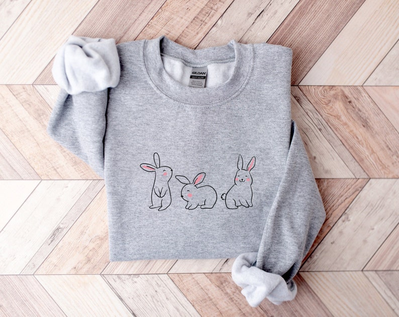 May include: A grey sweatshirt featuring a graphic of three bunnies in a row. The bunnies are drawn in a simple, cartoon style with pink noses and ears.