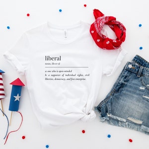 May include: White t-shirt with the definition of the word "liberal" printed on it. The definition is: "noun, lib-er-al a: one who is open-minded be a supporter of individual rights, civil liberties, democracy, and free enterprise."