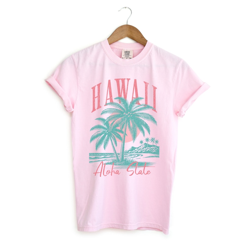 May include: Pink t-shirt with a graphic of palm trees and the text "Hawaii Aloha State" in a vintage style.