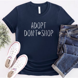 May include: A navy blue t-shirt with the text "Adopt Don't Shop" in white with a paw print symbol. The shirt is laid flat on a white surface with a pair of white sneakers and a pair of blue jeans.