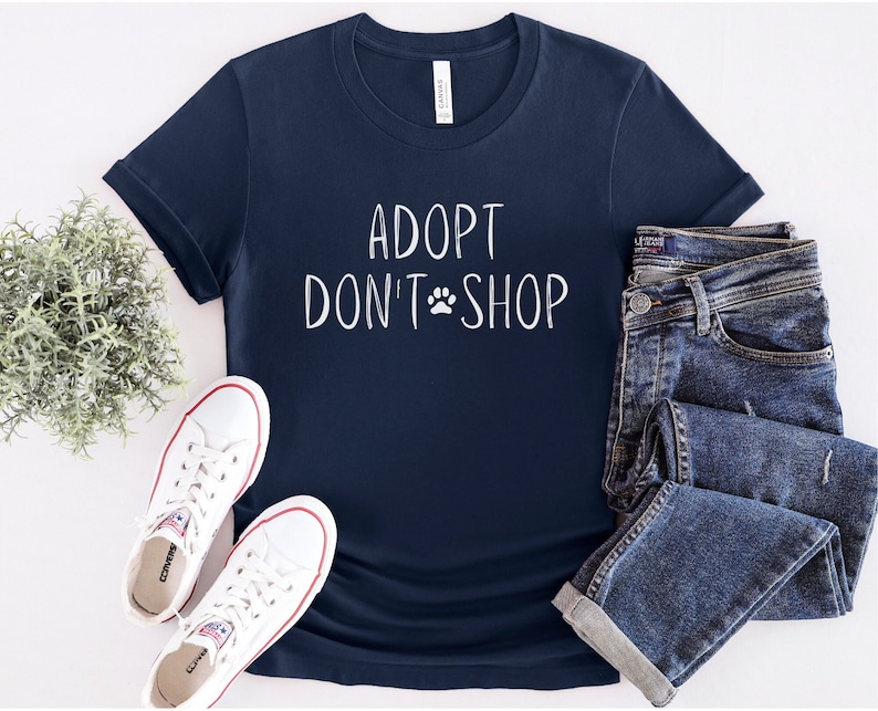 May include: A navy blue t-shirt with the text "Adopt Don't Shop" in white with a paw print symbol. The shirt is laid flat on a white surface with a pair of white sneakers and a pair of blue jeans.