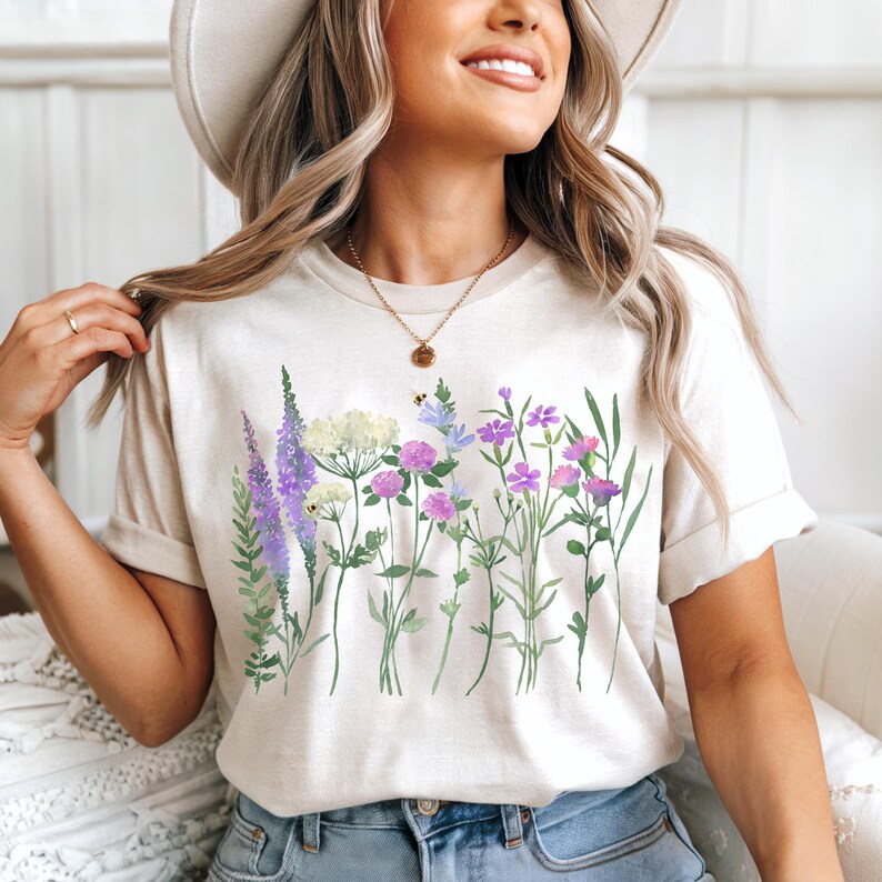 May include: Beige t-shirt featuring a watercolor floral design. The design includes purple, pink, and white flowers with green stems and leaves. The shirt has short sleeves and a crew neck.