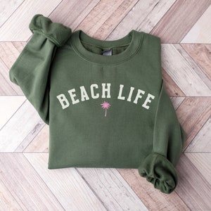 Pode incluir: Uma camisa de moletom verde com o texto "BEACH LIFE" em branco e um gr&aacute;fico de palmeira rosa.