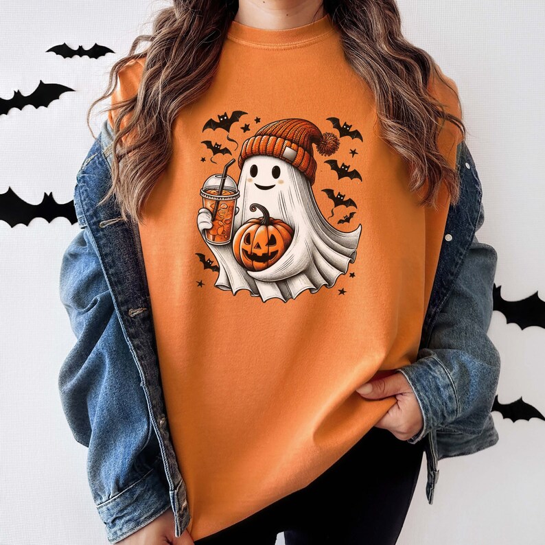 May include: Orange sweatshirt with a cute cartoon ghost wearing a hat and holding a pumpkin and a cup of coffee. The ghost is surrounded by bats and stars.
