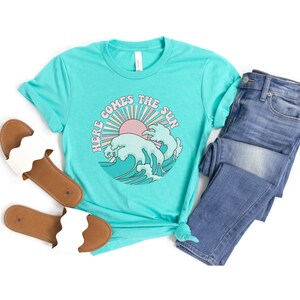 Op de afbeelding: Een turquoise blauw t-shirt met een retro afbeelding van een zon en golven. De afbeelding zegt "Here Comes The Sun".