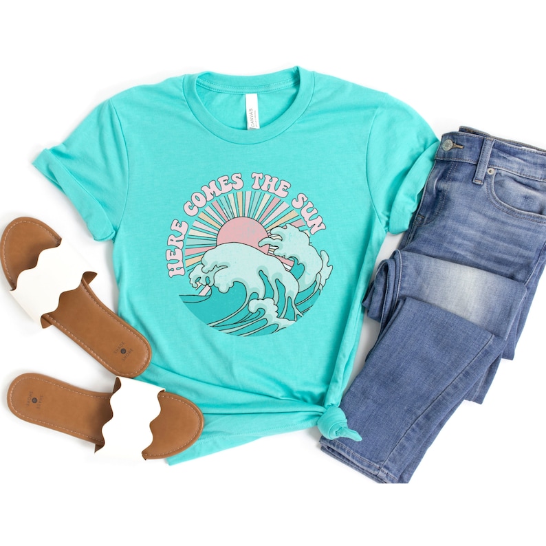 Op de afbeelding: Een turquoise blauw t-shirt met een retro afbeelding van een zon en golven. De afbeelding zegt "Here Comes The Sun".