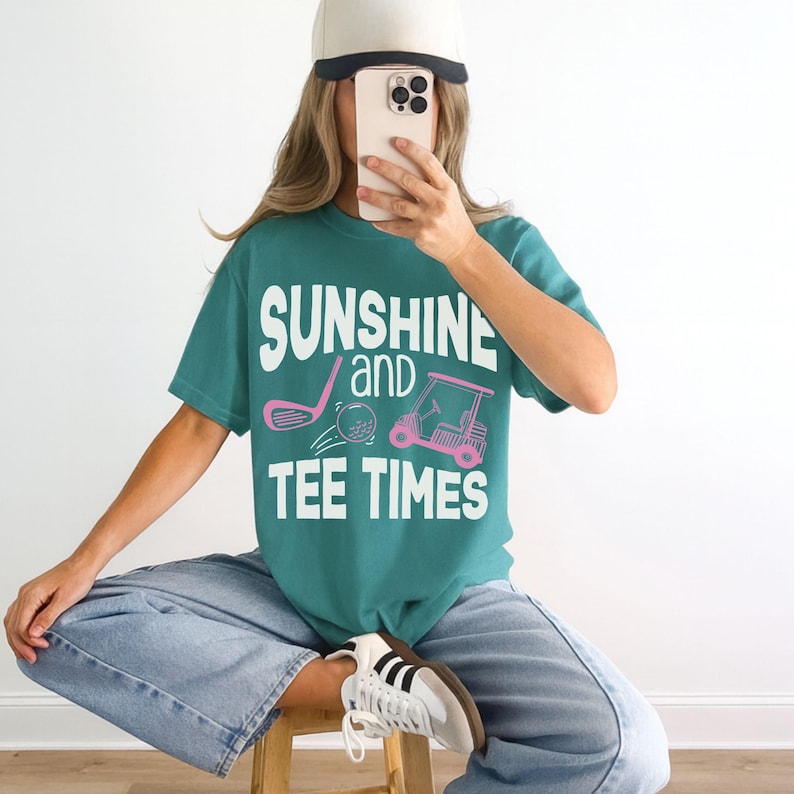以下が含まれることがあります： 「SUNSHINE and TEE TIMES」の白文字とピンク色のゴルフをテーマにしたグラフィックがプリントされた、ティール色のTシャツ。明るいブルーのジーンズと白いベースボールキャップを着用しています。