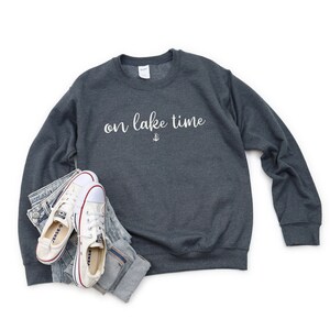 Puede incluir: Una sudadera gris oscuro con el texto "on lake time" en letras blancas y un peque&ntilde;o s&iacute;mbolo de ancla debajo del texto.