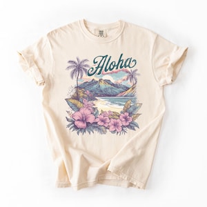 以下が含まれることがあります： トロピカルな風景デザインのクリーム色のTシャツ。グラフィックには、ターコイズ色のスクリプトで「Aloha」という言葉、ヤシの木、山、ビーチ、ピンクの花が含まれています。シャツは柔らかい素材で作られています。