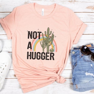 May include: A pink t-shirt with the text "NOT A HUGGER" in black with a rainbow and two cactus plants in green, brown, and pink.