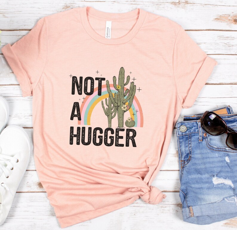 May include: A pink t-shirt with the text "NOT A HUGGER" in black with a rainbow and two cactus plants in green, brown, and pink.