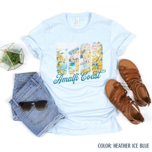 May include: Pale blue t-shirt with an Amalfi Coast graphic, showing lemons and coastal views. The words "Amalfi Coast" are in teal script. The shirt is styled with denim shorts, sunglasses, and brown sandals.