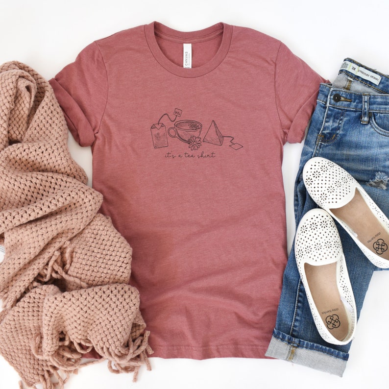 May include: A maroon t-shirt with a graphic of a teacup, tea bag, and tea infuser. The text "it's a tea shirt" is printed below the graphic.
