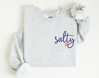 Könnte beinhalten: Ein grauer Pullover mit dem Wort "salty" in dunkelblauer Kursivschrift und einem rosa Seestern darunter.