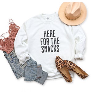 May include: White sweatshirt with the text "HERE FOR THE SNACKS" in black lettering. The sweatshirt is paired with blue jeans, a pink lace bralette, and leopard print flats.