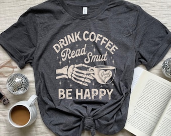 May include: A dark gray t-shirt with the text "Drink Coffee Read Smut Be Happy" printed on it. A skeleton hand is holding a coffee cup with a heart that says "Good girl" on it.