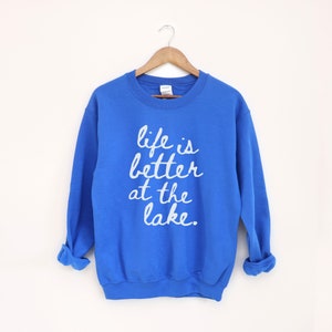 Op de afbeelding: Een koninklijk blauw sweatshirt met de tekst "life is better at the lake" in wit gedrukt op de voorkant.