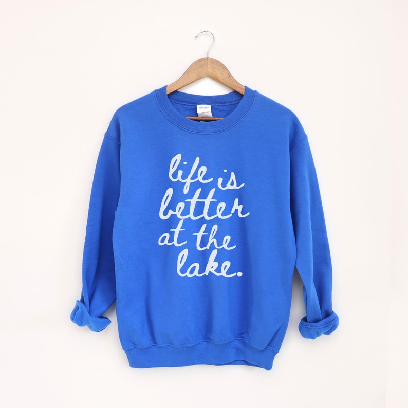 Op de afbeelding: Een koninklijk blauw sweatshirt met de tekst "life is better at the lake" in wit gedrukt op de voorkant.