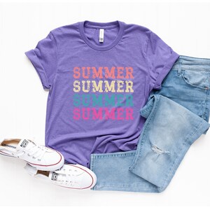 May include: A purple t-shirt with the word "SUMMER" printed four times in different colors. The shirt is paired with light blue jeans and white canvas sneakers with red accents. The items are arranged on a white surface.