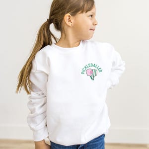 May include: White sweatshirt with a green and pink pickleball paddle graphic and the text "Pickleballer" on the front.