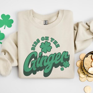 K&ouml;nnte beinhalten: Ein cremefarbenes Sweatshirt mit dem gr&uuml;nen Schriftzug "Dibs on the Ginger" und einem Kleeblatt-Design. Das Sweatshirt liegt gefaltet auf einer wei&szlig;en Oberfl&auml;che, mit Kleeblatt-Ausschnitten und einer Schale mit Goldm&uuml;nzen.