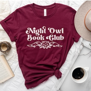 May include: A maroon t-shirt with the text "Night Owl Book Club" in white, featuring a stylized owl with wings spread out.