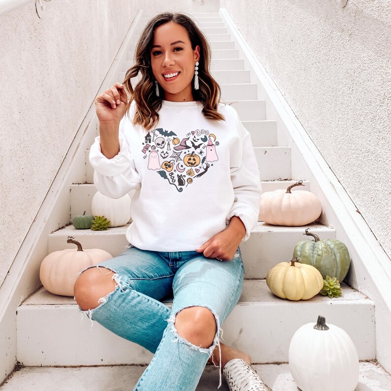 Op de afbeelding: Een wit sweatshirt met een roze en zwart hartontwerp met verschillende Halloween-iconen, waaronder geesten, pompoenen, vleermuizen en een heksenhoed. Het sweatshirt wordt gedragen door een persoon die op een witte trap zit met pompoenen op de treden.