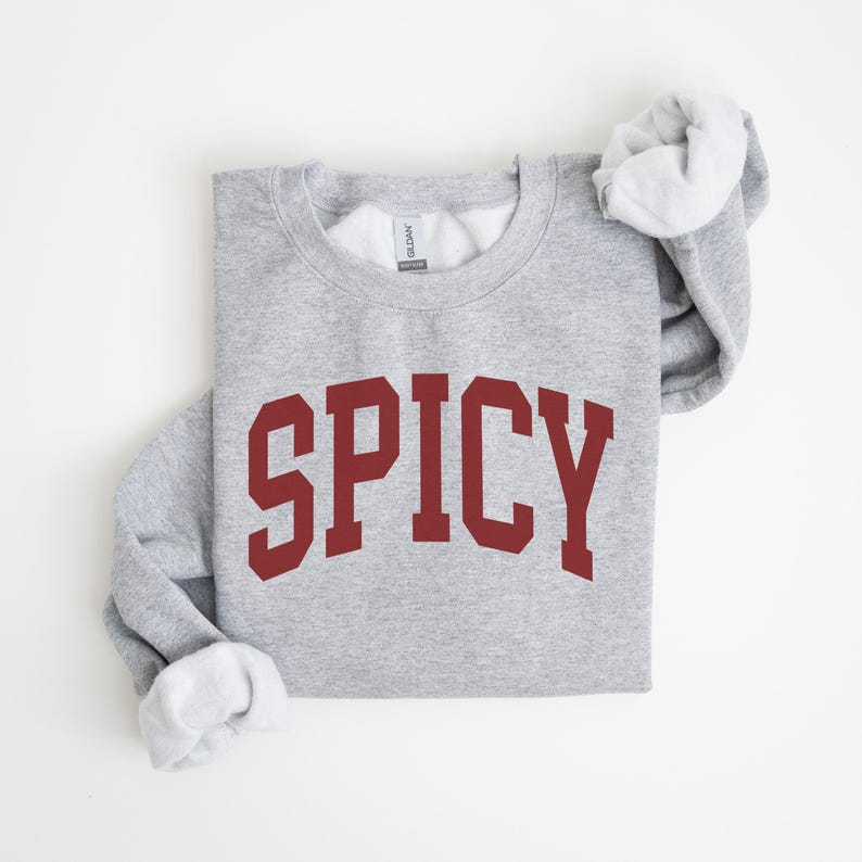 K&ouml;nnte beinhalten: Ein grauer Pullover mit dem Wort "SPICY" in Rotbraun auf der Vorderseite.