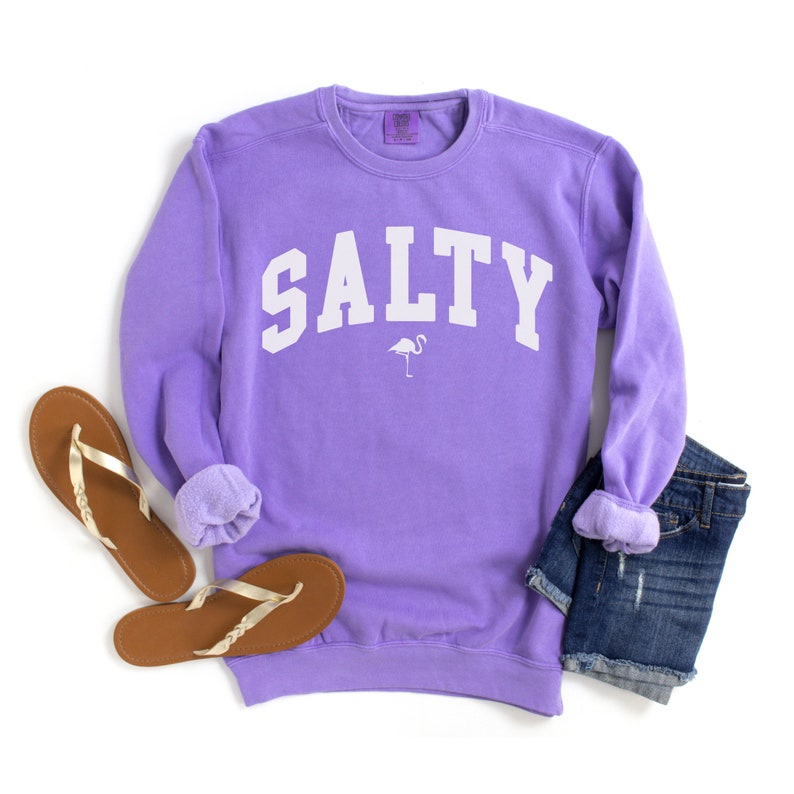 Op de afbeelding: Een lavendelkleurig sweatshirt met het woord "SALTY" in wit op de voorkant. Een klein wit silhouet van een flamingo bevindt zich onder het woord. Het sweatshirt ligt op een witte achtergrond met een paar bruine slippers en een blauwe spijkerbroek.