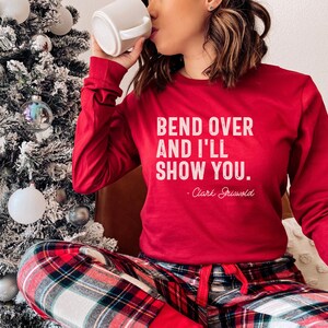 May include: Red long-sleeved shirt with the text "Bend over and I'll show you. - Clark Griswold" in white. The person is wearing red and black checked pyjama bottoms.