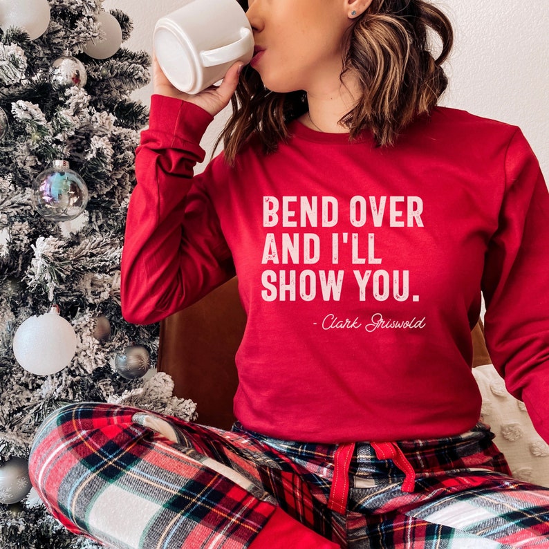 May include: Red long-sleeved shirt with the text "Bend over and I'll show you. - Clark Griswold" in white. The person is wearing red and black checked pyjama bottoms.