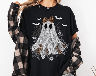 May include: Black t-shirt with a white ghost design, featuring a lace-like pattern, a brown bow, and floral details. The design includes bats and spiderwebs, ideal for Halloween. The shirt is a comfortable, casual wear.