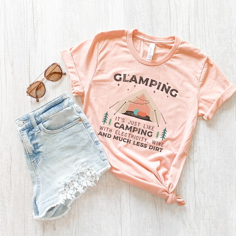 May include: A pink t-shirt with the text "GLAMPING" and "IT'S JUST LIKE CAMPING WITH ELECTRICITY, WINE AND MUCH LESS DIRT". The shirt features a cartoon illustration of a tent with fairy lights and a campfire.