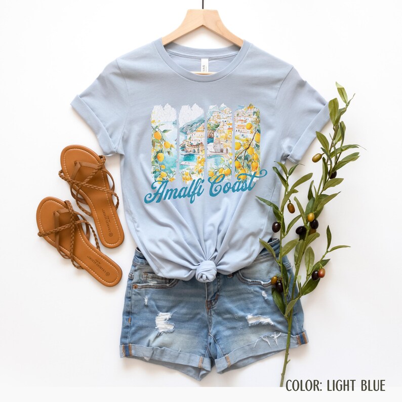 May include: Pale blue t-shirt featuring an Amalfi Coast design with coastal illustrations and lemons. The shirt is styled with denim shorts and brown sandals. A sprig of olives is placed on the side.
