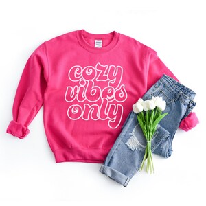 May include: A pink sweatshirt with the text "cozy vibes only" in white outlined letters. The sweatshirt is laid out on a white surface with a pair of blue jeans and a bouquet of white flowers.