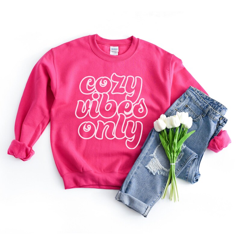 May include: A pink sweatshirt with the text "cozy vibes only" in white outlined letters. The sweatshirt is laid out on a white surface with a pair of blue jeans and a bouquet of white flowers.