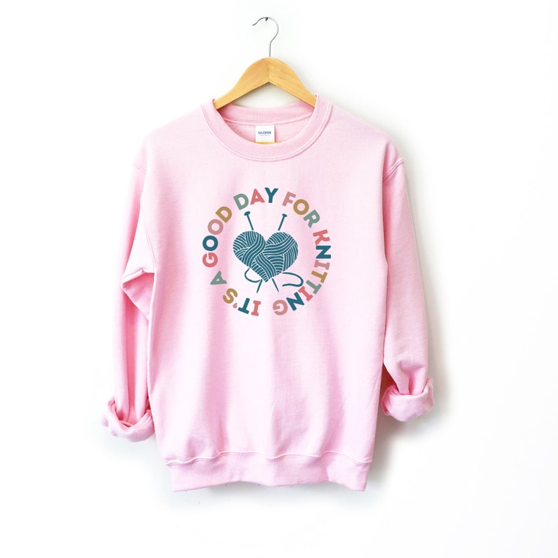K&ouml;nnte beinhalten: Rosafarbener Pullover mit einem grafischen Design, auf dem "Es ist ein guter Tag zum Stricken" steht, mit einem herzf&ouml;rmigen Wollkn&auml;uel und Stricknadeln.