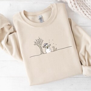 Op de afbeelding: Beige crewneck sweatshirt met een grillige wintersc&egrave;ne. Een sneeuwpop met een hoed en sjaal staat bij een kale boom, met een hond. Sneeuwvlokken vallen. Het ontwerp is in zwart en wit.