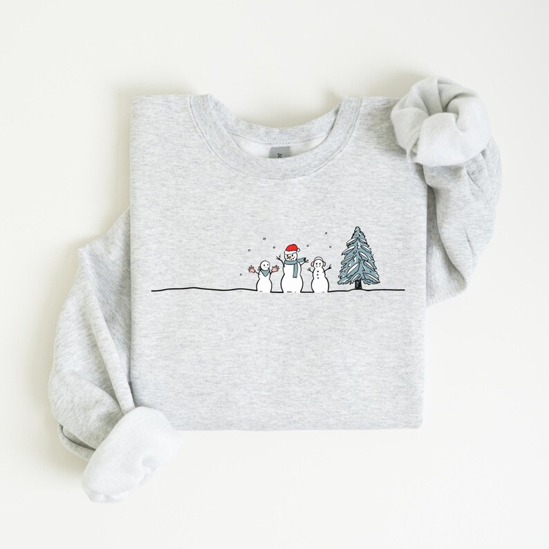 Op de afbeelding: Een lichtgrijs sweatshirt met een winterontwerp. De illustratie toont drie sneeuwpoppen, een kerstboom en vallende sneeuw. De sneeuwpoppen dragen een rode hoed, een blauwe sjaal en een blauwe hoed. Het sweatshirt is opgevouwen.