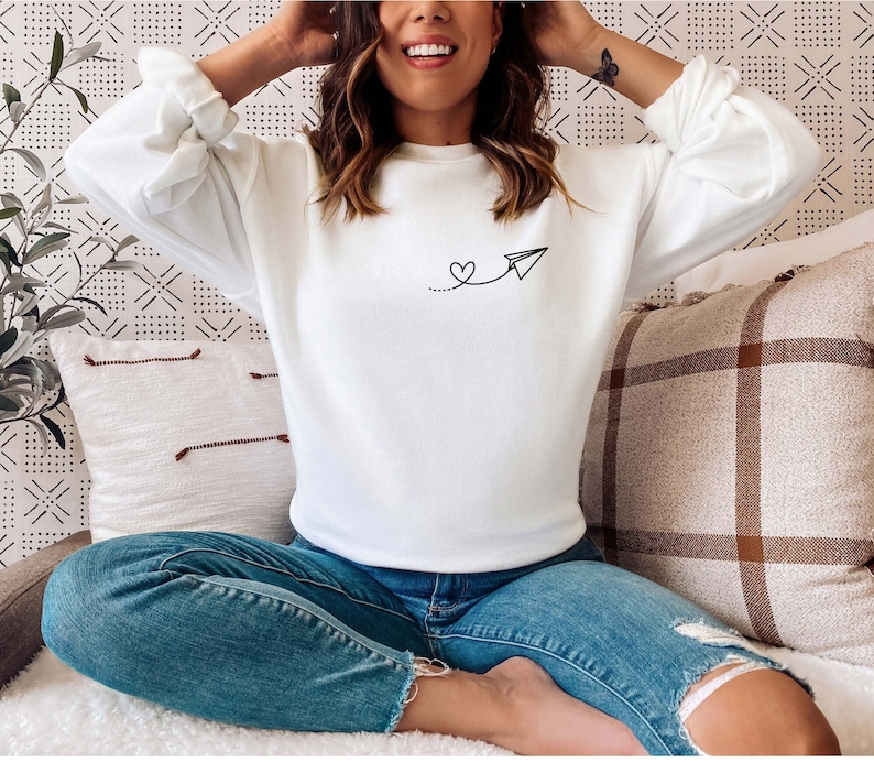 Op de afbeelding: Wit sweatshirt met een simpel zwart ontwerp van een papieren vliegtuig met een hartje aan de staart. Het model draagt een blauwe jeans.