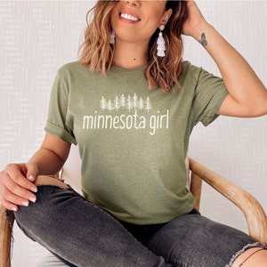 May include: A green t-shirt with a white graphic of pine trees and the text "minnesota girl".