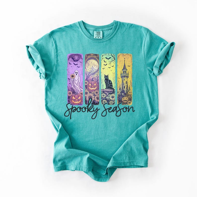 May include: Teal-colored t-shirt featuring a Halloween-themed graphic. The graphic includes a ghost, jack-o'-lanterns, a black cat, and a spooky tower. The words "Spooky Season" are written in a cursive font below the graphic.