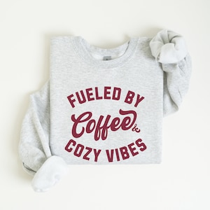 May include: A light grey sweatshirt featuring the burgundy text "Fueled by Coffee & Cozy Vibes". The sweatshirt is folded, with a rolled-up sleeve, and is presented on a white background. The crew neck sweatshirt is a casual style.