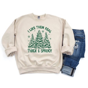 May include: A beige sweatshirt with a green graphic of three snow-covered pine trees. The text "I like them real thick & sprucy" is printed on the sweatshirt.