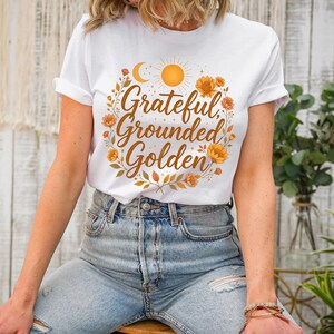 Pu&ograve; includere: T-shirt bianca con le parole "Grateful, Grounded, Golden" in un design circolare con un sole, una luna e accenti floreali. La maglietta &egrave; abbinata a jeans di denim chiaro.